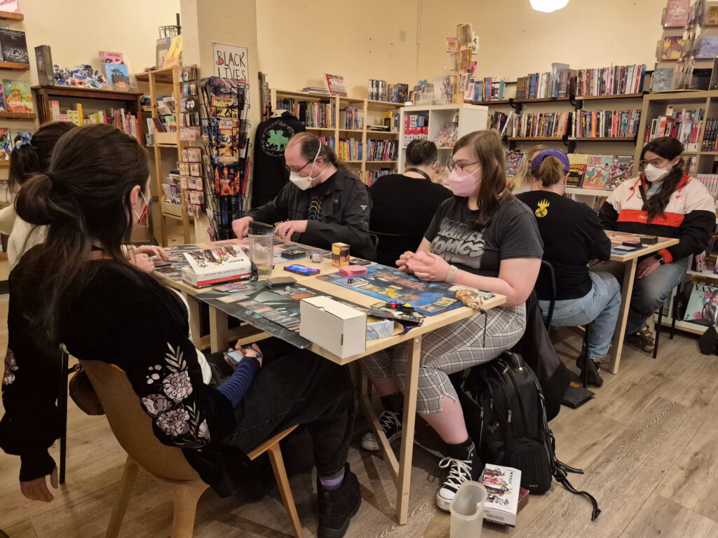Photo of 8 people at tables playing a trading card game. They are all wearing masks. Bookshelves are in the background.