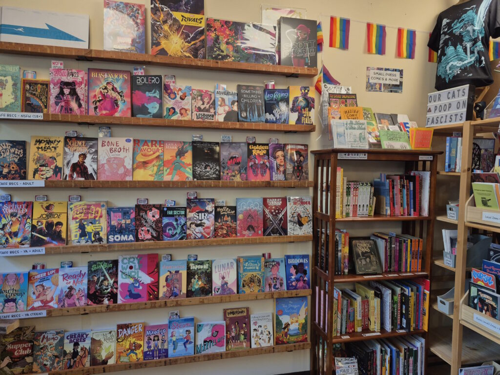 Photo of interior of bookstore with some bookshelves as well as 6 rows of books on the wall facing out. Some LGBTQ+ pride flags are seen in the corner.